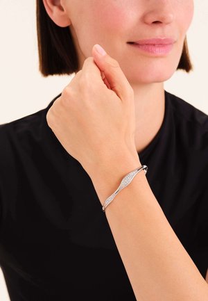 Bracciale d'argento con design ondulato, caratterizzato da una finitura liscia e pietre trasparenti incastonate, indossato su un polso con tono di pelle chiara.