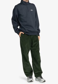 Granatowa bluza z półsuchem z małym nadrukiem na piersi, zestawiona z zielonymi spodniami sztruksowymi. Całość ma luźny krój i jest uzupełniona szarymi trampkami.