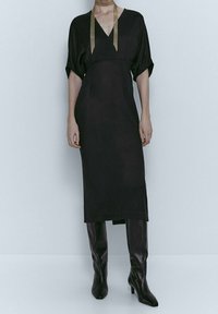 Sort kjole med V-udskæring, korte dolman-ærmer og lige snit. Har guldsløjfeaccents ved halsen og ankelhøje sorte støvler.