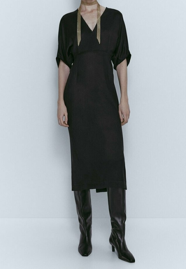 Sort kjole med V-udskæring, korte dolman-ærmer og lige snit. Har guldsløjfeaccents ved halsen og ankelhøje sorte støvler.