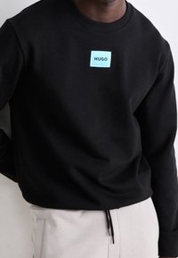 Sudadera negra con cuello redondo, con un parche de logo rectangular azul que muestra "HUGO" en el pecho. Textura suave.