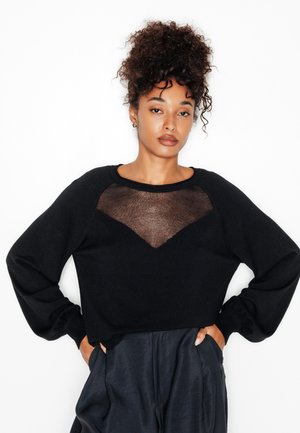 Donna con capelli ricci raccolti in uno chignon alto, indossa un maglione nero con pannello trasparente a forma di V e pantaloni neri, in piedi con le mani nelle tasche.