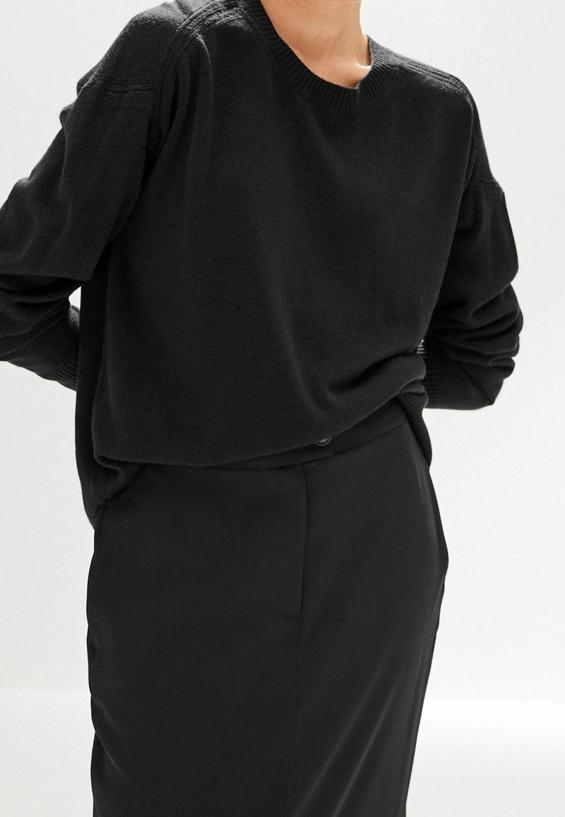 Personne portant un pull noir uni à manches longues légèrement rentré dans un pantalon noir taille haute sur fond blanc.