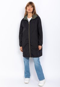 Cappotto impermeabile nero con uno strato interno verde, chiusura con cerniera e maniche lunghe. Indossato con jeans azzurri chiari e sneaker bianche.