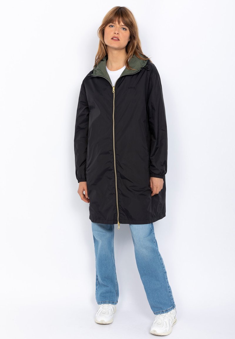 Manteau noir imperméable avec une doublure intérieure verte, fermeture éclair et manches longues. Porté avec un jean bleu clair et des baskets blanches.