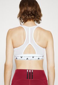 Biała sportowa biustonosz z designem racerback i wycięciem. Posiada teksturowany pas z czarnym logo Adidas i paskami.