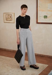 Black short-sleeve top, light gray wide-leg trousers, black platform sandals, holding a textured black tote bag. Minimalist design, neutral tones.