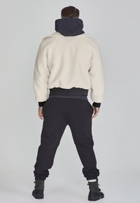 Jachetă pufoasă de crem cu glugă închisă, manșete elastice și pantaloni de trening negri. Jacheta are un design scurt și prezintă o suprafață texturată.