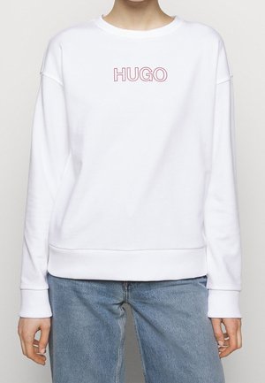 Vit sweatshirt med rundad halsringning och ribbade ärmslut. "HUGO"-logotyp i rosa i mitten. Matchas med ljusblå jeans.