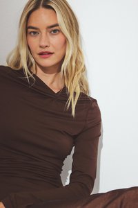 Blonde ženska z razpuščenimi lasmi, oblečena v temno rjavo majico z dolgimi rokavi, pozira ob enobarvni beli steni.