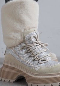 Bottes épaisses avec un revers en fausse fourrure beige, des accents en strass argentés et une semelle à plateforme texturée, présentant un design à lacets sur le devant.