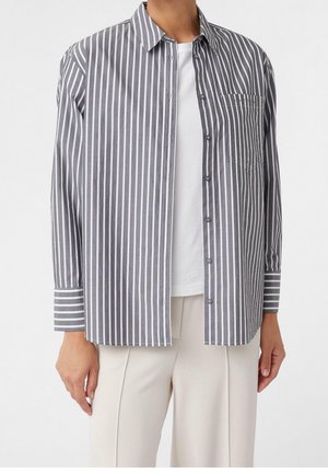 Person wearing a gray and white vertical striped button-up shirt over a white top with beige pants, standing against a plain white background.