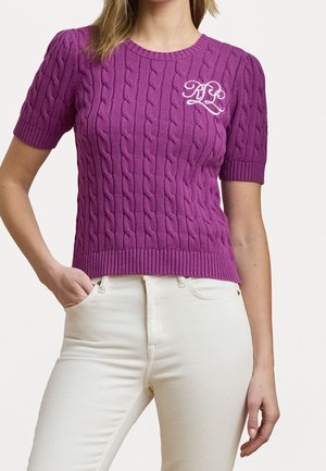 Femme portant un pull violet à manches courtes en maille torsadée avec des initiales blanches brodées et un pantalon écru taille haute, une main dans la poche.