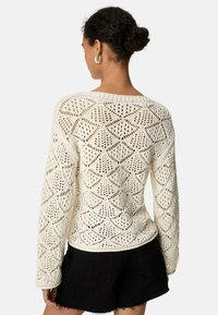 Vit, virkad tröja med lång ärm, med diamantmönstrade utskärningar. Lätt material, avslappnad passform, som bärs över svarta shorts.