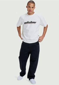 Vit t-shirt med "Quiksilver"-logotyp i svart och grått, kombinerad med löst sittande svarta byxor och vita sneakers.