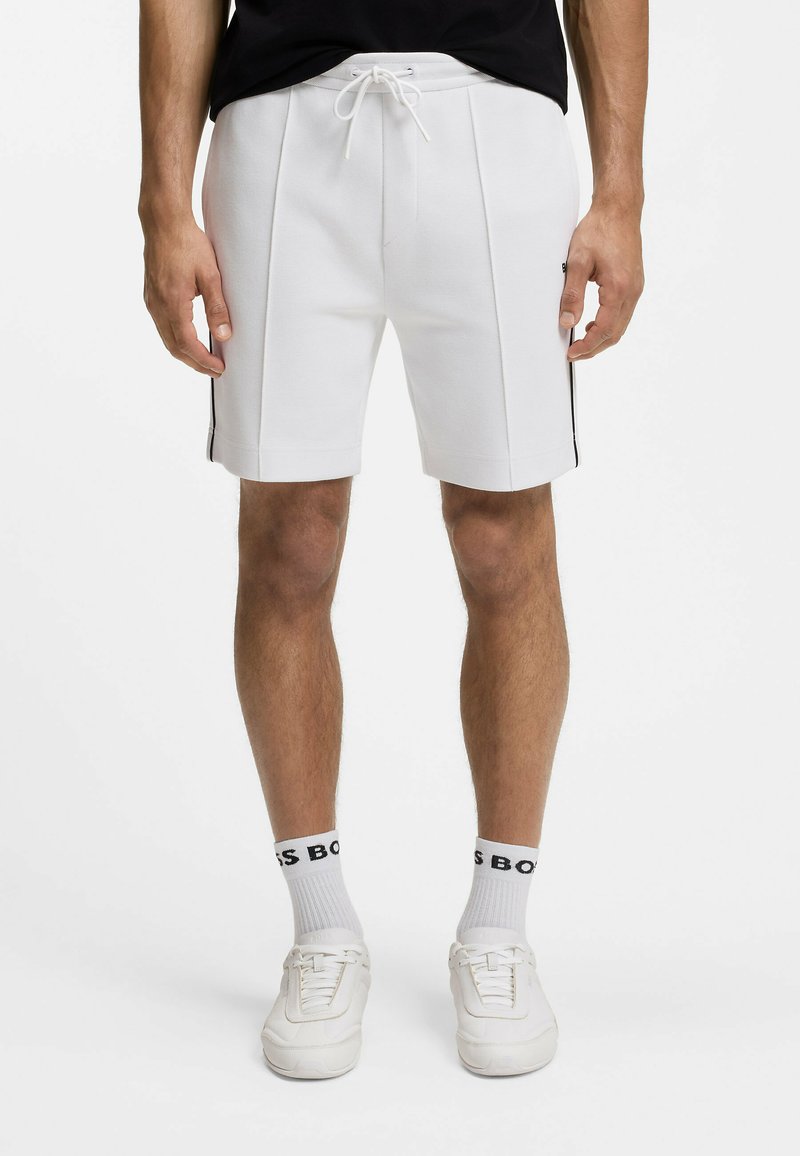 Hombre vistiendo pantalones cortos blancos con cordón y franjas negras en los lados, calcetas blancas con letras negras y zapatillas blancas, de pie frente a un fondo blanco.