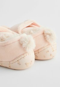 Chaussons roses avec des pompons moelleux, en tissu doux, et une semelle à motifs floraux. La forme arrondie et les bords cousus mettent en valeur les détails du design.