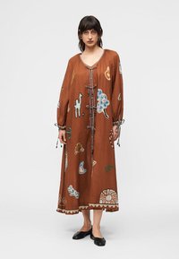 Γυναίκα που φοράει ένα μακρύ, φαρδύ καφέ φόρεμα με πολύχρωμα αφηρημένα και ανθισμένα σχέδια, δεμένο μπροστά, και μαύρα επίπεδα παπούτσια, όρθια μπροστά σε απλό φόντο.