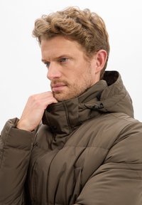 Brązowa pikowana kurtka z wysokim kołnierzem, zatrzaskami i widocznym przeszyciem. Materiał wydaje się gładki, z kieszeniami z przodu.