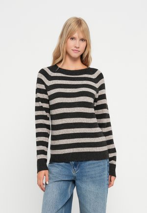 Vrouw staand in een zwart-grijs horizontaal gestreepte trui en lichtblauwe spijkerbroek tegen een effen witte achtergrond.