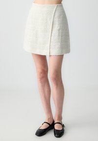 Femme portant une mini-jupe texturée crème et des ballerines noires, se tenant contre un fond clair uni.
