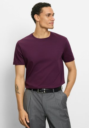 Mann trägt ein pflaumenfarbenes T-Shirt mit Rundhalsausschnitt, das in graue, plissierte Hosen mit schwarzem Gürtel gesteckt ist, steht mit einer Hand in der Tasche und blickt seitlich.