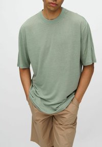Camiseta de mangas cortas de corte sencillo en tejido verde claro, ligeramente texturizado, con cuello redondo y fit relajado, combinada con pantalones cortos beige.