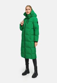 Groene pufferjas met een hoge kraag en capuchon, voorzien van een tweezijdige rits en zijzakken. Draagt over zwarte leggings en chunky zwarte laarzen.