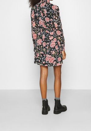 Blumenkleid in Schwarz mit hohem Ausschnitt, langen Ärmeln und gerafftem Bund. Hat pinke und blaue Blumen. Getragen mit schwarzen Stiefeln.