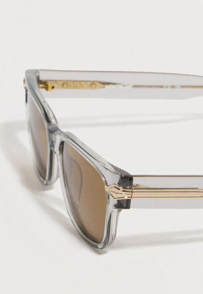 Διαφανή γυαλιά ηλίου με γκρι σκελετό, καφέ φακούς και χρυσές λεπτομέρειες στα πλευρικά μέρη, εμφανιζόμενα από κοντινή πλάγια όψη.