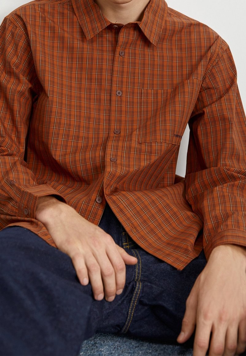 Homme portant une chemise à carreaux orange à boutons et un jean bleu foncé, assis avec les mains reposant sur les cuisses.