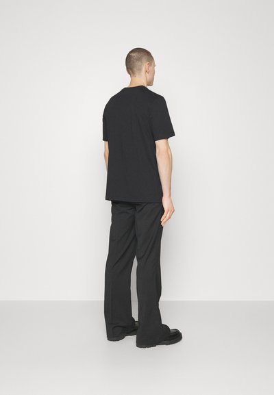 Black cotton t-shirt with short sleeves and a round neckline, worn with wide-leg black trousers, viewed from the back. Smooth texture.