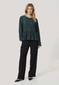 Türkisfarbenes Langarmoberteil mit strukturiertem Oberteil und Peplum-Saum, kombiniert mit schwarzen, weit geschnittenen Hosen und spitzen Schuhen.
