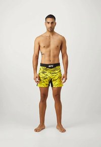  Άντρας που στέκεται ξυπόλητος, χωρίς μπλούζα, φορώντας κίτρινα και μαύρα σορτς UFC με γραφικό μοτίβο, μπροστά σε απλό ανοιχτό φόντο.