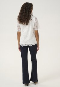 White lace top with short sleeves and scalloped edges, paired with dark flared jeans. The lace features floral and geometric patterns.
