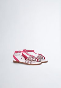 Vaaleanpunaiset sandaalit, joissa on nauhamallinen muotoilu. Yläosa on valmistettu teksturoidusta kankaasta, ja sandaaleissa on säädettävä nilkkahihna sekä matala pohja, jossa on vaaleanruskea viimeistely.