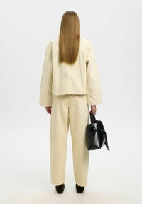 Kremowy bawełniany zestaw z krótką kurtką o pudełkowym kroju i szerokimi spodniami. Modelka trzyma czarną skórzaną torebkę, do której uwiązana jest apaszka z wzorem.