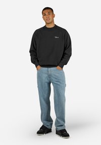 Sort sweatshirt med rund halsudskæring og ribstrikkede manchetter, båret med lyseblå jeans. Sweatshirten har et lille mærkelogo på brystet.