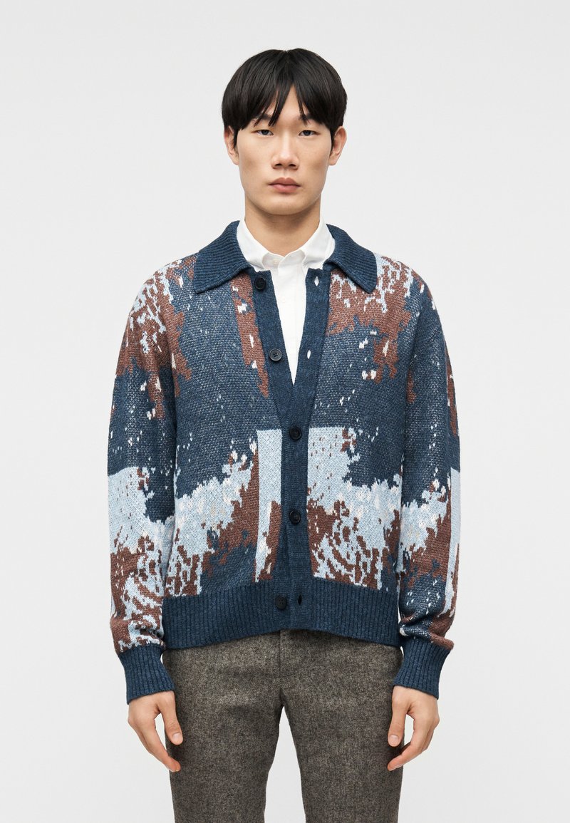 Νέος άνδρας που φοράει ζακέτα με μπλε, λευκό και καφέ αφηρημένα σχέδια πάνω από λευκό πουκάμισο και καφέ υφασμένα παντελόνια, στέκεται μπροστά σε απλό φόντο.