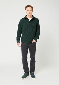 Giovane uomo in piedi con una mano in tasca, che indossa una camicia a maniche corte a bottone di colore verde scuro con motivo, jeans neri e sneakers verdi su sfondo bianco.