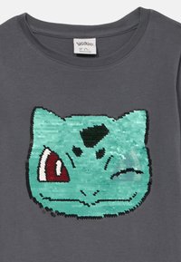 Szara koszulka z dużym wzorem Bulbasaura ozdobionym zielonymi cekinami, z czerwonymi i czarnymi akcentami. Okrągły dekolt, materiał bawełniany.