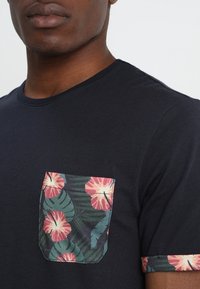 Camiseta oscura de color azul marino con un bolsillo y puños de manga con un patrón floral tropical. Tela suave con colores contrastantes: rosa, verde y fondos oscuros.