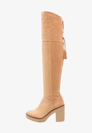 Botte haute en daim beige, talon bloc, bout arrondi et détail de nœud à pampilles en haut à l'arrière.