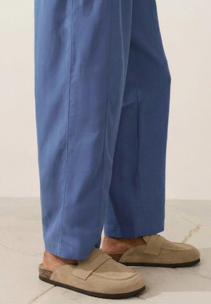 Mocassins beige en daim sans lacets portés avec un pantalon droit bleu sur un sol carrelé clair.