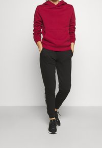 Punainen puuvillainen huppari, jossa edessä tasku, yhdistettynä mustiin collegehousuihin, joissa valkoinen sivuraita. Mustat urheilukengät täydentävät asun.