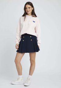 Un sweat à capuche rose avec un logo bleu, une mini-jupe plissée marine avec des boutons dorés, et des baskets montantes blanches avec des détails en lacets.