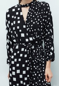 Schwarzes Wickelkleid mit weißen Quadratmustern, langen Ärmeln, einer taillierten Taille und einem Schlüssel-Ausschnitt. Weicher Stoff mit strukturiertem Finish.