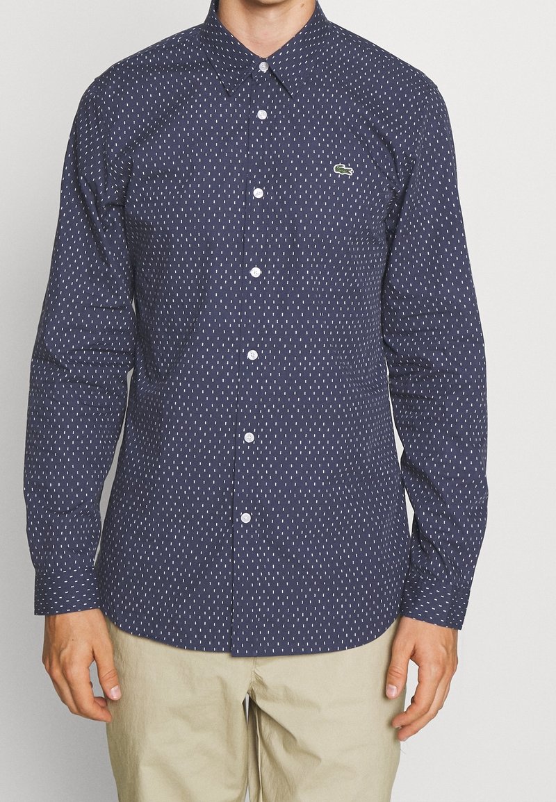 Homme portant une chemise bleu marine à manches longues boutonnée avec un motif à pois blancs et un pantalon beige, debout devant un fond uni.