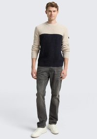 Strickpullover mit einem oben cremefarbenen und unten dunkelblauen Abschnitt, versehen mit strukturierten Mustern. Kombiniert mit grauen Jeans und weißen Sneakers.