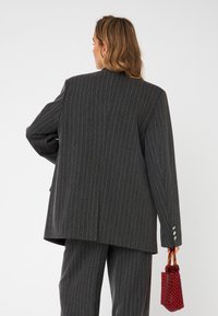 Blazer grigio a righe con design oversize, dotato di schiena ventilata e dettagli con bottoni argentati. Abbinato a una piccola borsa a tracolla di perline rosse.